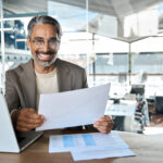 Homme au bureau qui lit un document papier à coté de son laptop.